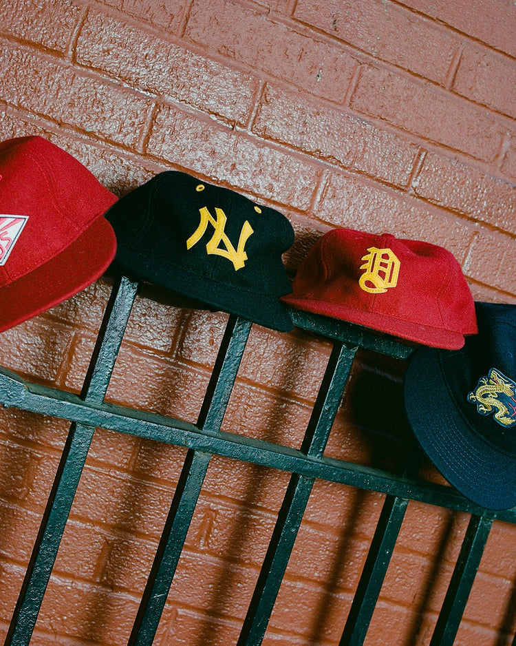 Four baseball caps, including the Ebbets Field Taiyo Cubs 1928 Authentic Wool Ballcap, are displayed on a black metal fence in front of a red brick wall.