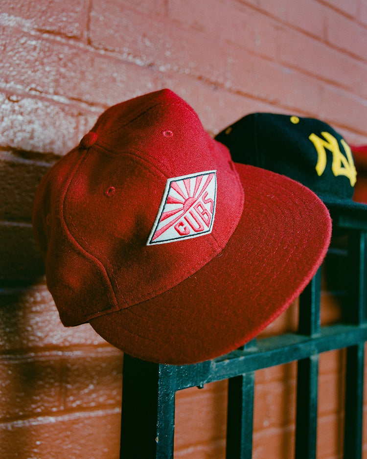 A red Taiyo Cubs 1928 Authentic Wool Ballcap by Ebbets Field with a white and red logo sits on a black metal fence, while a blurred dark cap with yellow lettering appears in the background against a brick wall.