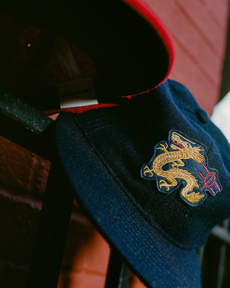A close-up of the Chunichi Dragons 1969 Wool Ballcap by Ebbets Field, with a yellow dragon and red basketball logo, resting on a black metal railing near a red brick wall.