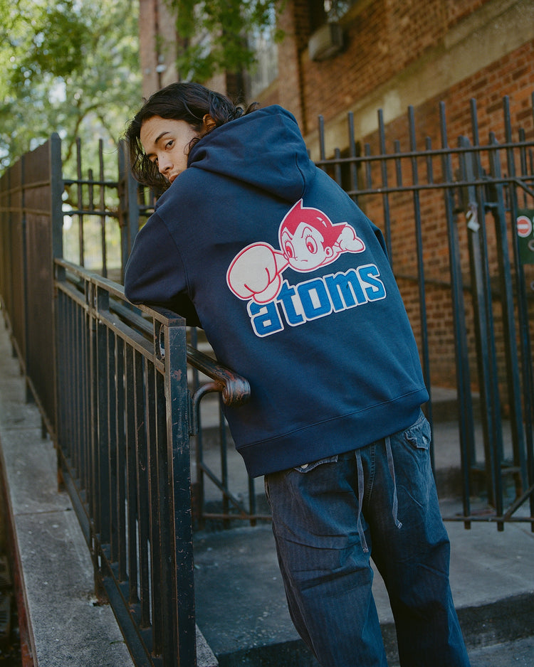 Someone in a navy Sankei Atoms Heavyweight Cotton Terry Full-Zip Hoodie by Ebbets Field, with a large cartoon graphic and “atoms” on the back, leans on a metal railing outside near a brick building.