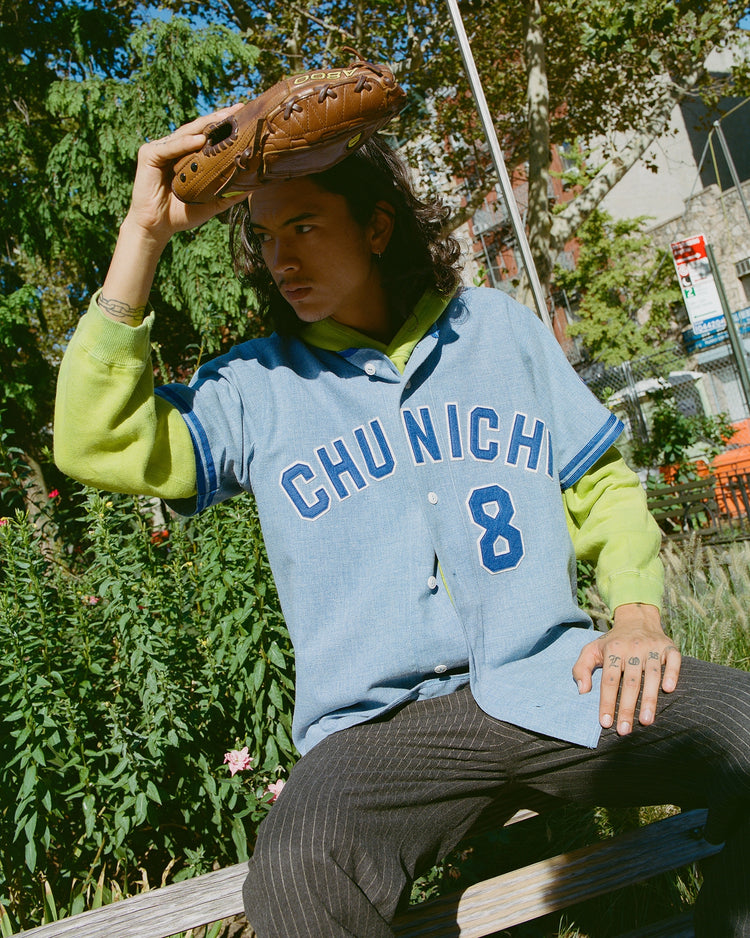 A young person with long hair sits on a bench outside, wearing the Chunichi Dragons 1969 Authentic Wool Road Jersey by Ebbets Field over a lime-green hoodie, holding a leather baseball glove as sunlight filters through trees.