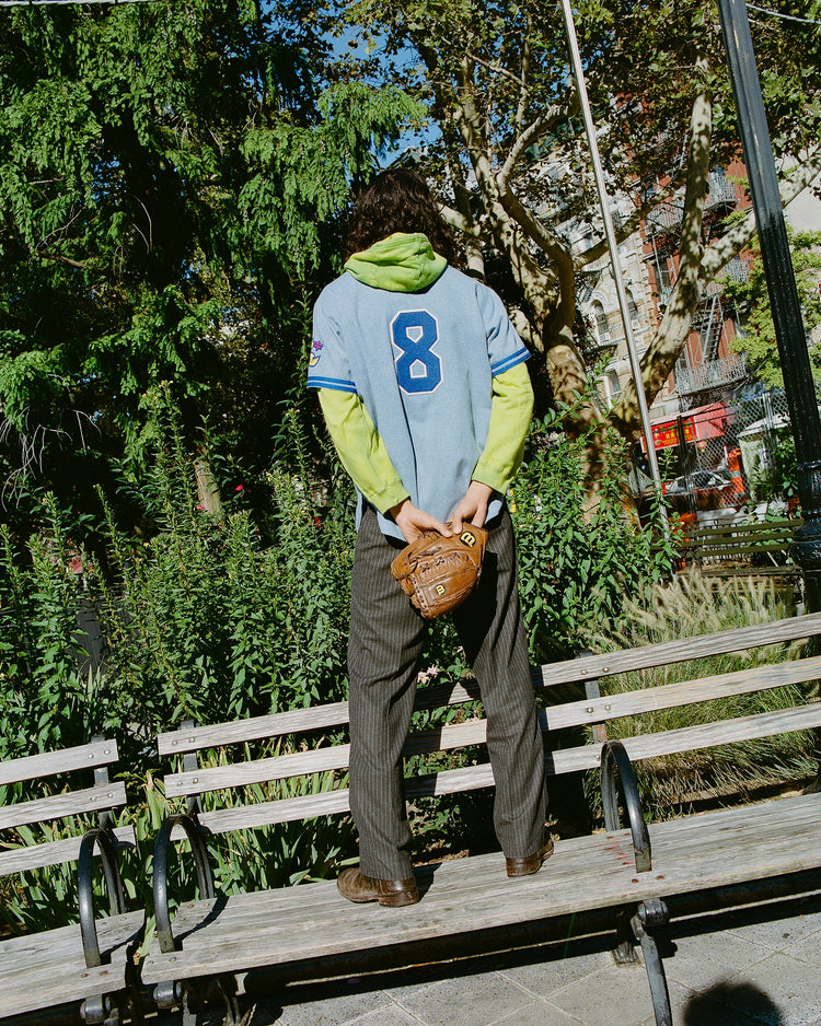 Wearing an Ebbets Field Chunichi Dragons 1969 Authentic Wool Road Jersey, a person with the number 8 stands on a park bench, holding a baseball glove behind their back amid lush greenery.