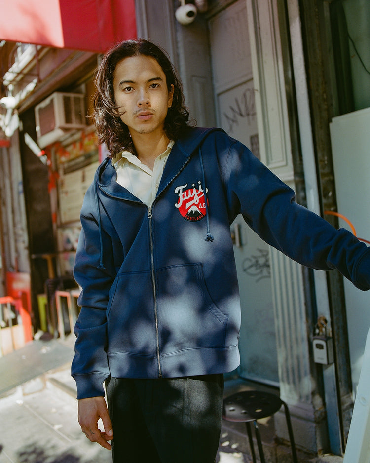A person with long dark hair wears the Fuji Athletic Club Heavyweight Cotton Terry Full-Zip Hoodie by Ebbets Field, standing outdoors in a sunlit urban area with graffiti and chairs in the background.