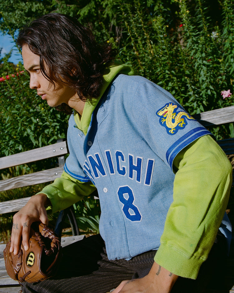 Someone wearing the Chunichi Dragons 1969 Authentic Wool Road Jersey by Ebbets Field with CHUNICHI and number 8 sits on a bench in a green hoodie, holding a baseball glove. Green plants and flowers fill the background.