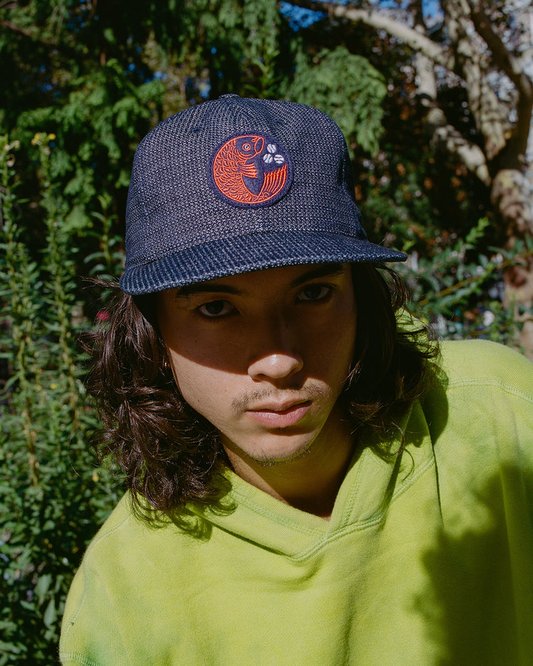 A person with long hair wears the Ebbets Field Hiroshima Carp Japanese Indigo Cotton Dobby Ballcap and a light green sweatshirt outdoors amid greenery and sunlight.