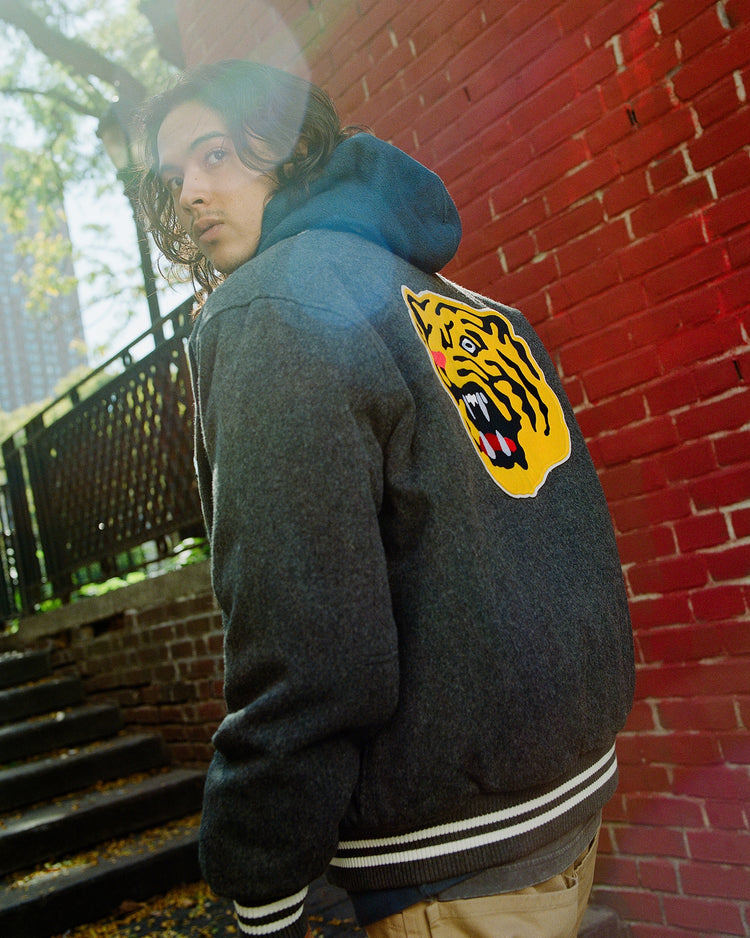 Wearing the Ebbets Field Osaka Tigers All-Wool Varsity Jacket featuring a bold tiger face patch, a person stands by red brick steps, glancing back as sunlight streams through the trees above.