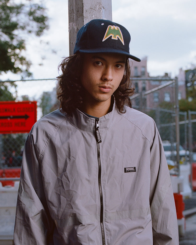 A young person with long hair wears a gray jacket and the Ebbets Field Nankai Hawks 1950 Authentic Wool Ballcap, featuring a yellow-green logo, standing outdoors near construction fencing and red signs.