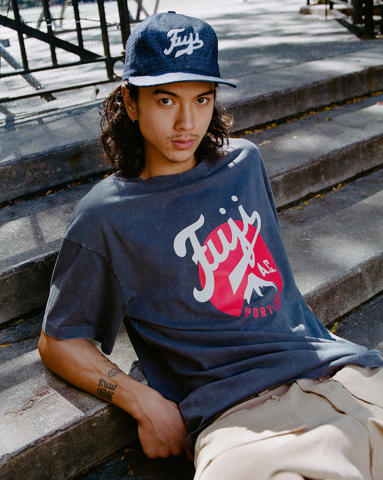 A person with long hair and a mustache sits outdoors wearing a navy cap and the Ebbets Field Fuji Athletic Club Pigment-Dyed Short Sleeve Graphic T-Shirt, looking relaxed at the camera.