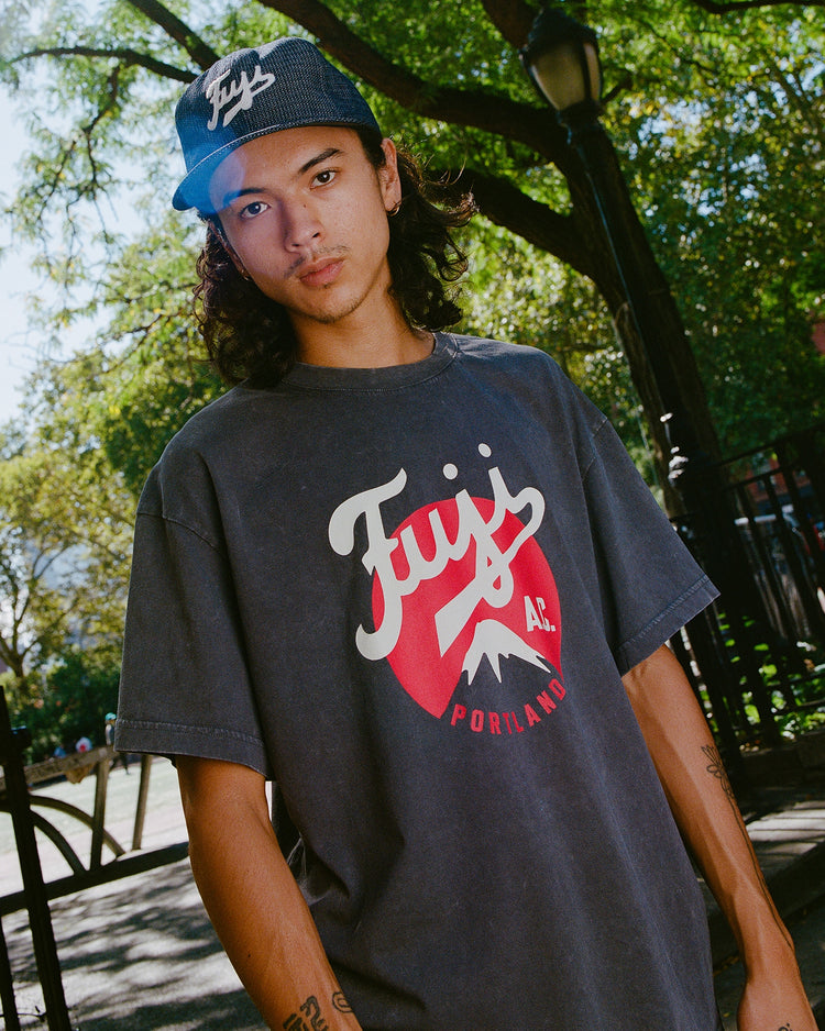 A young person with long hair wears an Ebbets Field Fuji Athletic Club Pigment-Dyed Short Sleeve Graphic T-Shirt and matching hat, standing outdoors by a green park and black metal fence.