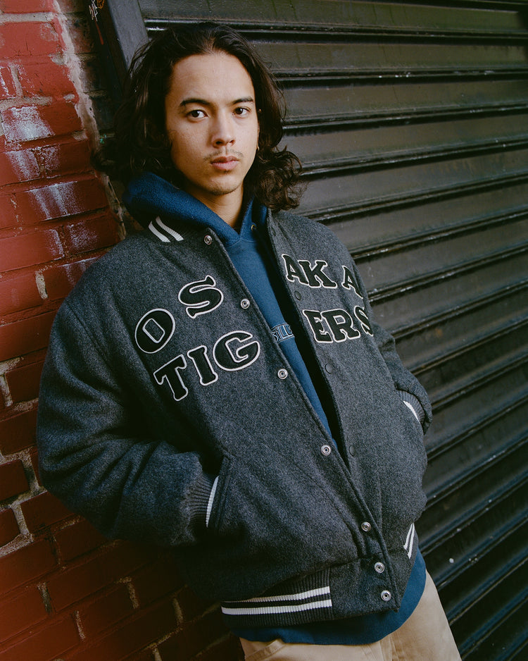 A person with long dark hair wears the Osaka Tigers All-Wool Varsity Jacket by Ebbets Field and a blue hoodie, standing against a brick wall and metal shutter, looking at the camera with hands in pockets.