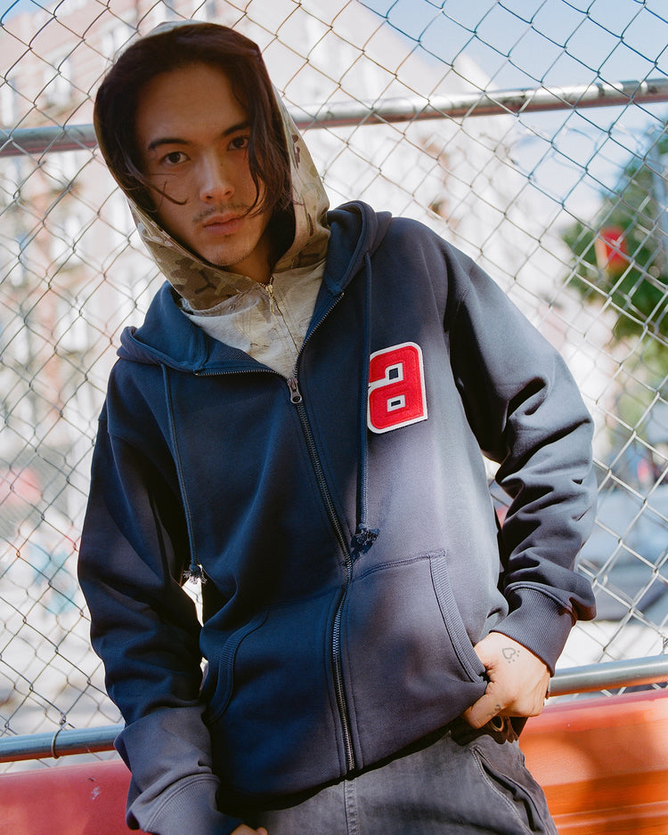 A person with long hair stands by a chain-link fence, wearing a patterned shirt under the Ebbets Field Sankei Atoms Heavyweight Cotton Terry Full-Zip Hoodie, featuring a red letter A patch.