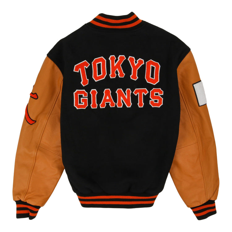 The Ebbets Field Tokyo Giants DNA Varsity Jacket features a black body, tan leather sleeves, TOKYO GIANTS in bold red and white on the back, black collar/cuffs/waistband with orange stripes, and sleeve patches.