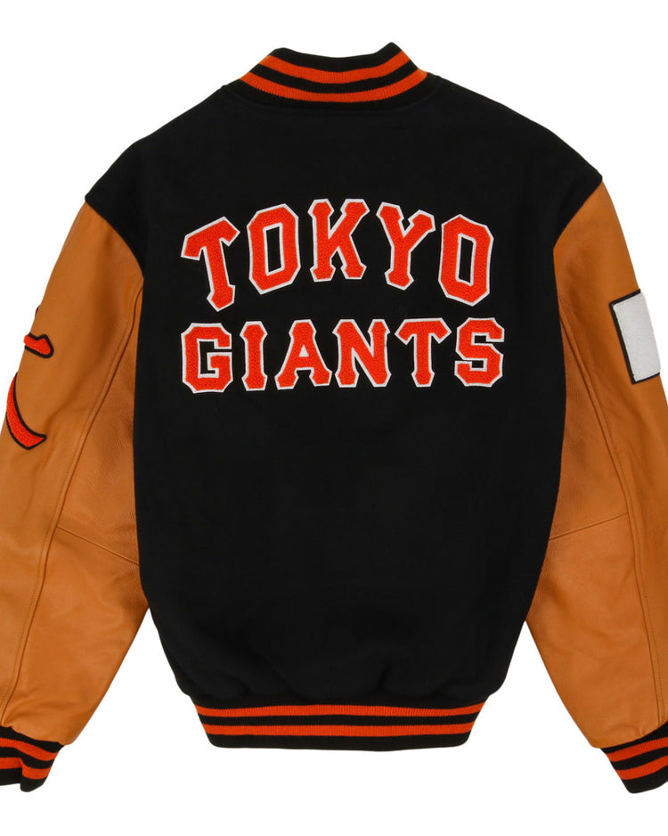 The Ebbets Field Tokyo Giants DNA Varsity Jacket features a black body, tan leather sleeves, TOKYO GIANTS in bold red and white on the back, black collar/cuffs/waistband with orange stripes, and sleeve patches.