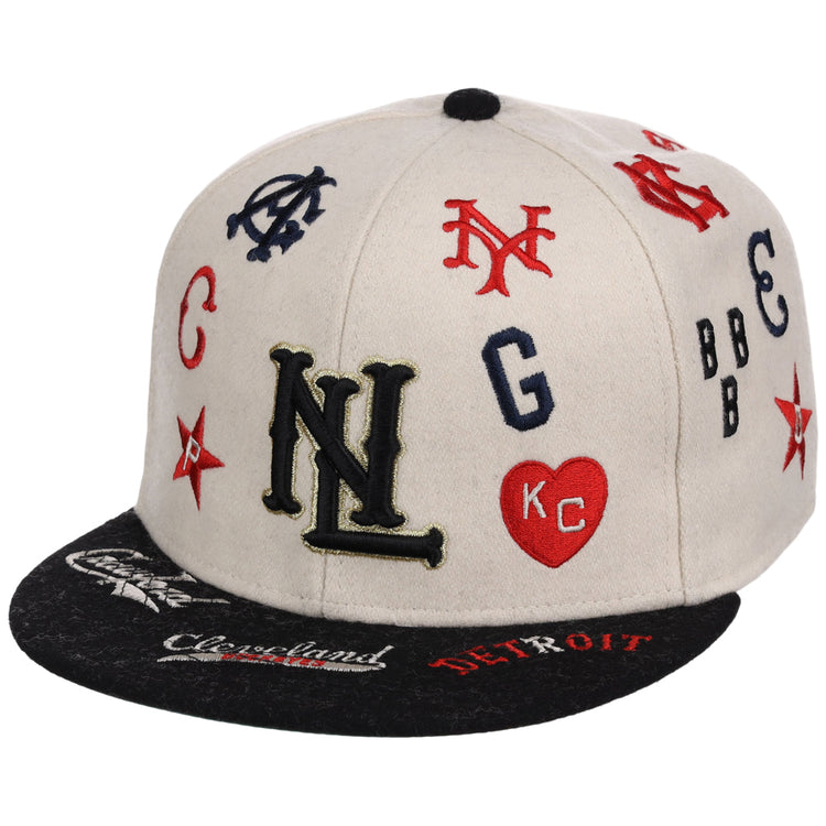 The Ebbets Field Negro League Baseball NLB Ballpark Fitted Ballcap features a beige crown, black brim, and colorful embroidered team logos and initials like NL, NY, KC in a heart, plus city names such as Cleveland and Detroit on the brim.