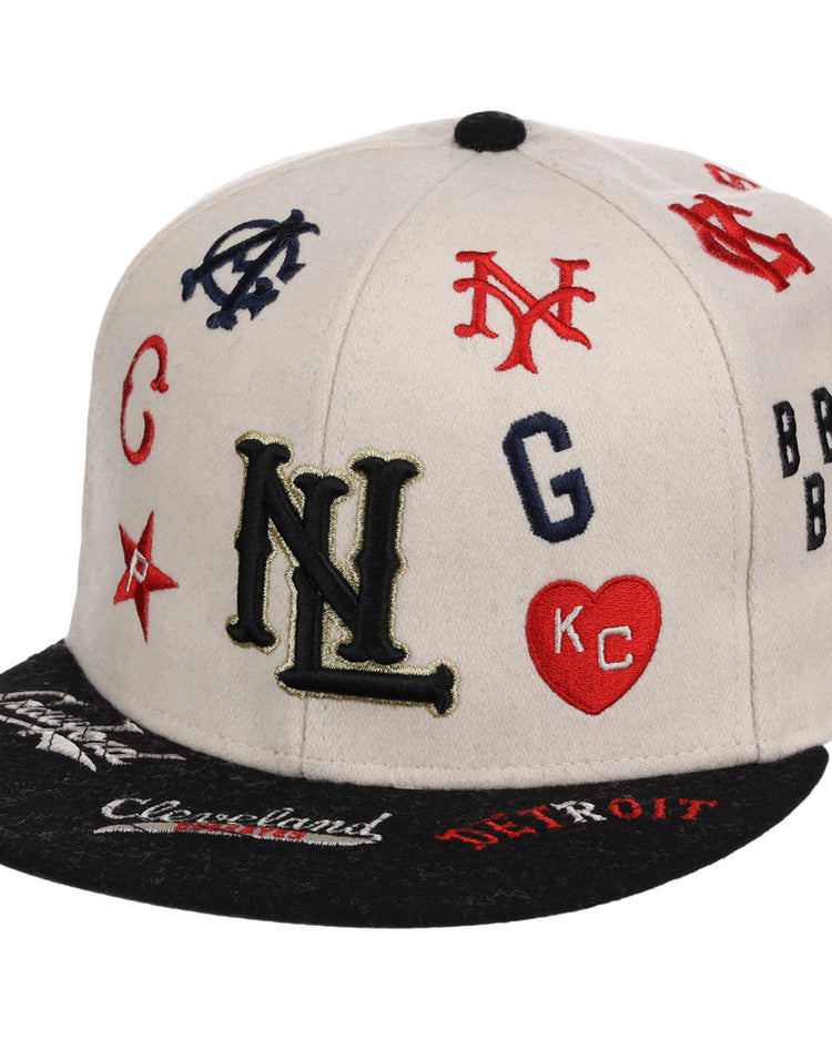 The Ebbets Field Negro League Baseball NLB Ballpark Fitted Ballcap features a beige crown, black brim, and colorful embroidered team logos and initials like NL, NY, KC in a heart, plus city names such as Cleveland and Detroit on the brim.