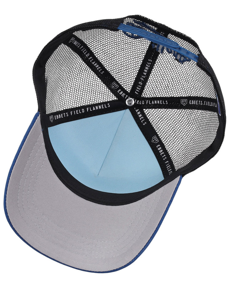 A top-down view of the Tulsa Drillers MiLB Trucker Ballcap by Ebbets Field shows its light blue crown, grey under-brim, black mesh panels, and interior branded black tape reading Ebbets Field Flannels.