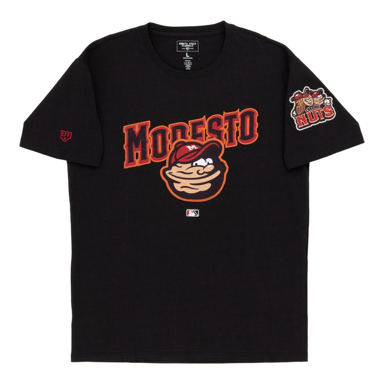 The Ebbets Field Modesto Nuts MiLB Fashion Cotton Short Sleeve Tee features a cartoon nut character, bold red MODESTO lettering, a chest logo, and a Nuts logo patch on the left sleeve.