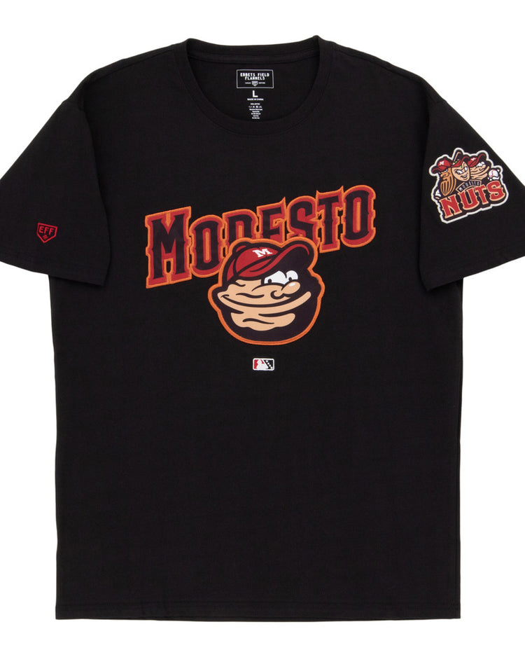 The Ebbets Field Modesto Nuts MiLB Fashion Cotton Short Sleeve Tee features a cartoon nut character, bold red MODESTO lettering, a chest logo, and a Nuts logo patch on the left sleeve.