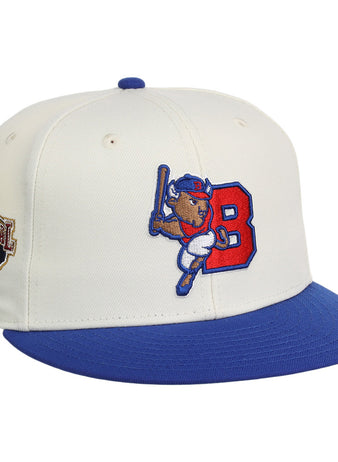 The Ebbets Field Buffalo Bisons MiLB Snapback Ballcap is a white cap with a blue brim, featuring a cartoon buffalo swinging a bat by a red 