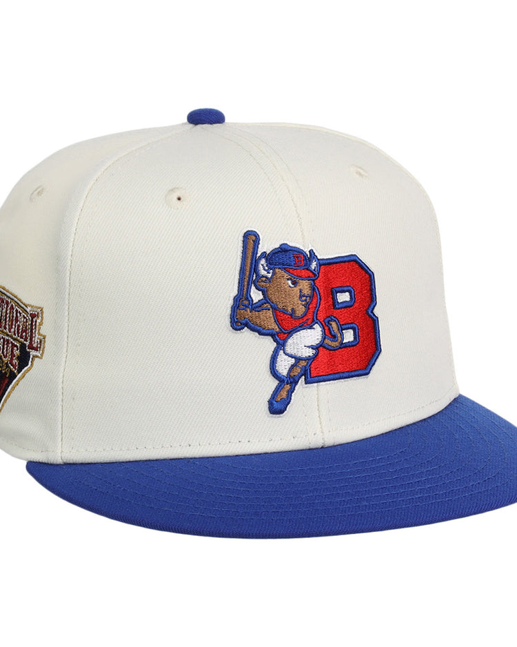 The Ebbets Field Buffalo Bisons MiLB Snapback Ballcap is a white cap with a blue brim, featuring a cartoon buffalo swinging a bat by a red 