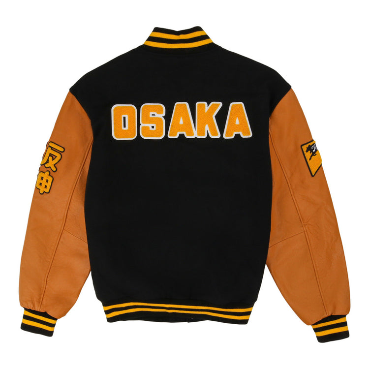 The Osaka Tigers DNA Varsity Jacket by Ebbets Field features a black and brown design, OSAKA in bold yellow and white on the back, yellow-striped cuffs/collar, and sleeve patches with Japanese characters.