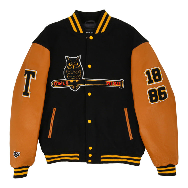 The Ebbets Field Topeka Owls MiLB Dna Varsity Jacket is black with tan leather sleeves, features an owl-on-bat patch, OWLS text, 1886 on the right sleeve, a large T on the left sleeve, and yellow striped collar and cuffs.
