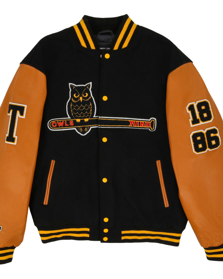 The Ebbets Field Topeka Owls MiLB Dna Varsity Jacket is black with tan leather sleeves, features an owl-on-bat patch, OWLS text, 1886 on the right sleeve, a large T on the left sleeve, and yellow striped collar and cuffs.