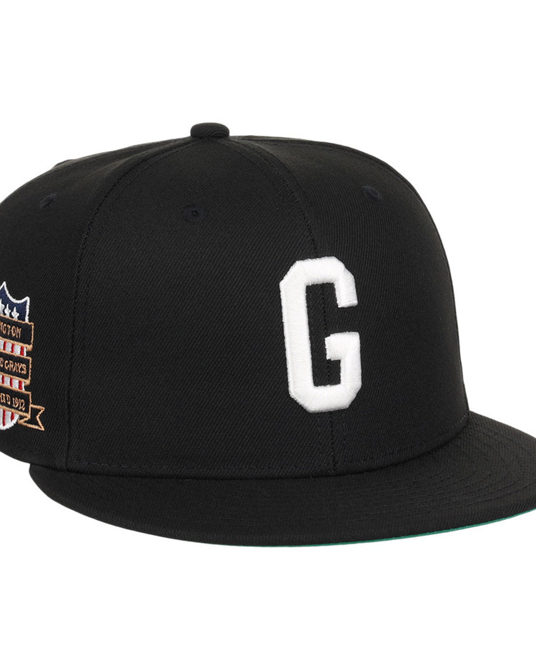 The Ebbets Field Homestead Grays NLB Tc Fitted Ballcap is a black cap with a white embroidered “G” on the front and a colorful patch featuring text and stars on the left side.