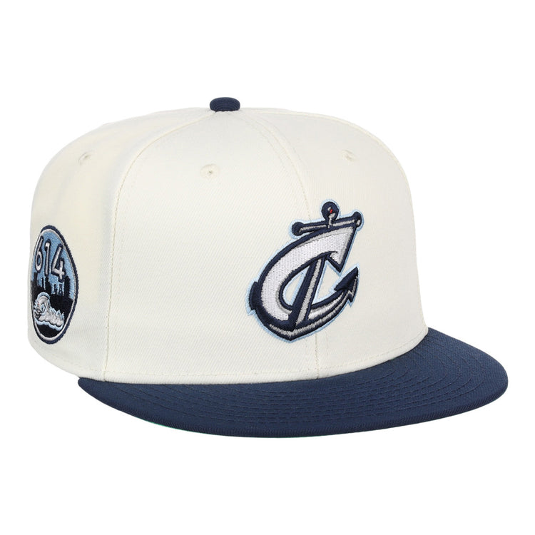 The Ebbets Field Columbus Clippers MiLB Snapback Ballcap is a white hat with a navy blue brim, featuring a stylized anchor and C logo on the front and a circular 614 cityscape patch on the side.