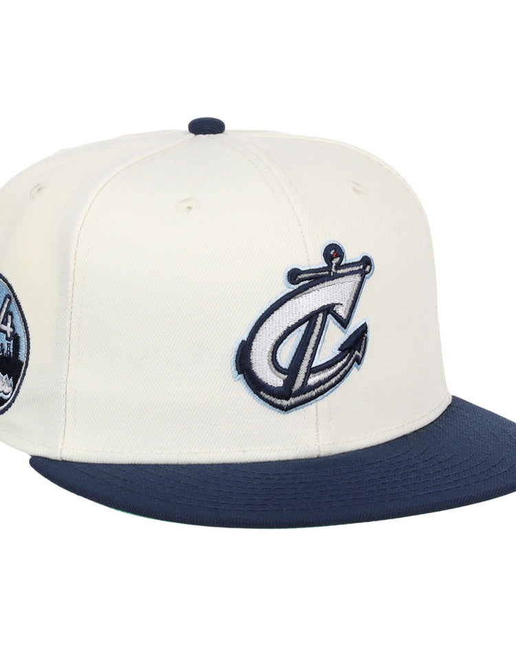The Ebbets Field Columbus Clippers MiLB Snapback Ballcap is a white hat with a navy blue brim, featuring a stylized anchor and C logo on the front and a circular 614 cityscape patch on the side.