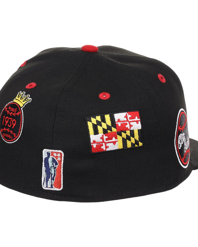 The Baltimore Elite Giants NLB Varsity Fitted Ballcap by Ebbets Field is a black cap featuring red, yellow, and white embroidered patches: a Maryland flag, a basketball-themed logo, and a person holding a basketball.