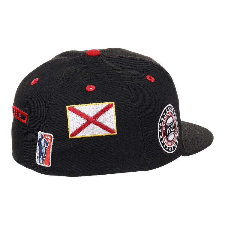 The Ebbets Field Birmingham Black Barons NLB Varsity Fitted Ballcap is a black cap with a flat brim, featuring patch details like a red and white flag, a 1924 circular logo, and a rectangular patch with a silhouetted figure.