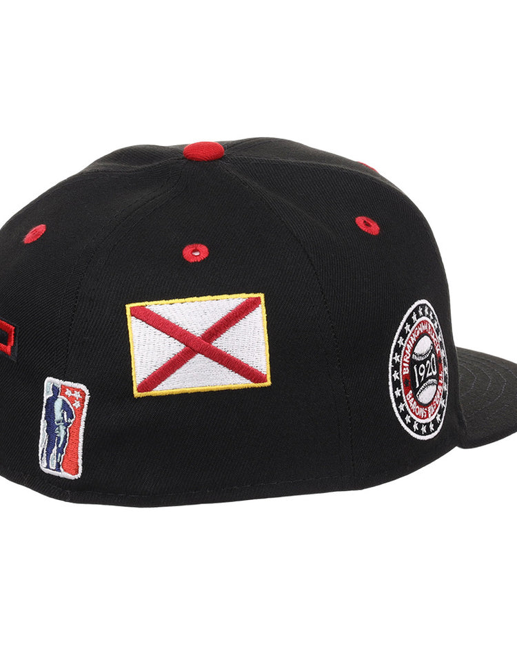 The Ebbets Field Birmingham Black Barons NLB Varsity Fitted Ballcap is a black cap with a flat brim, featuring patch details like a red and white flag, a 1924 circular logo, and a rectangular patch with a silhouetted figure.