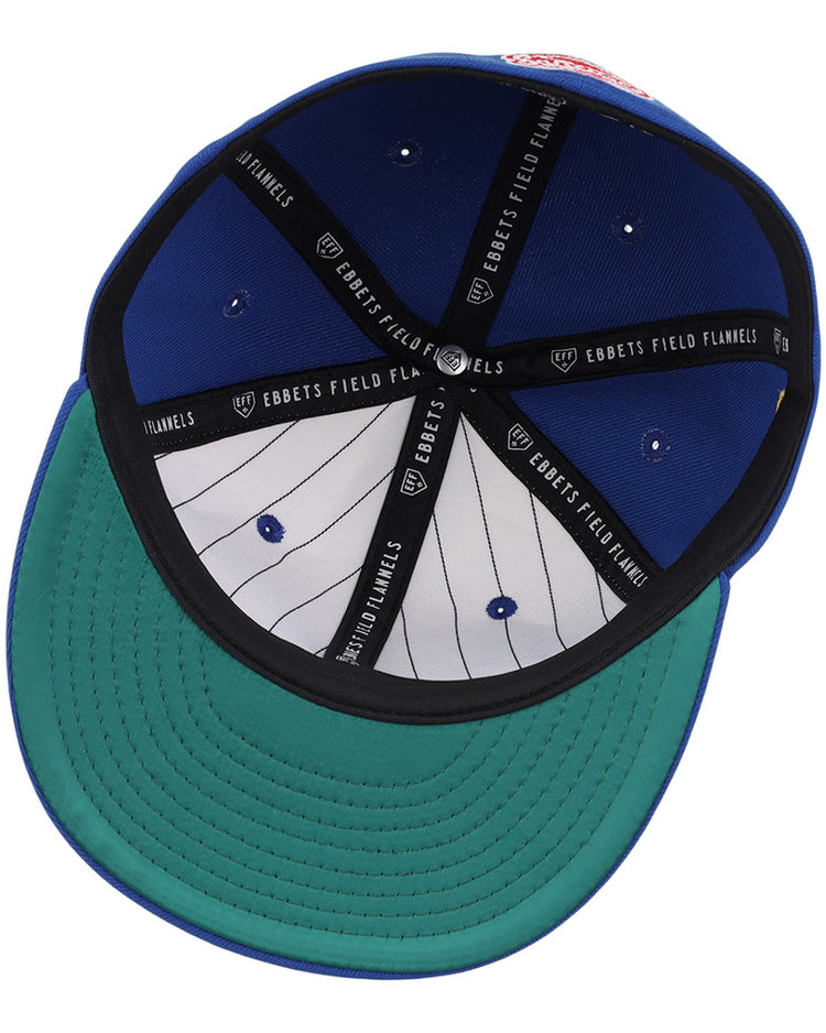 The image shows the inside of the Santurce Cangrejeros Wordmark Chainstitch Fitted Ballcap by Ebbets Field—a blue cap with a green brim, black Ebbets Field bands, and a white pinstriped lining with ventilation eyelets.
