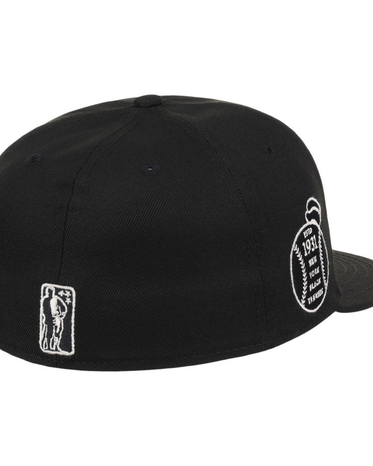 The Ebbets Field New York Black Yankees NLB Black/White Fitted Ballcap is shown from the back, with white embroidered patches—a small figure on the left and a football-shaped design with text on the right.