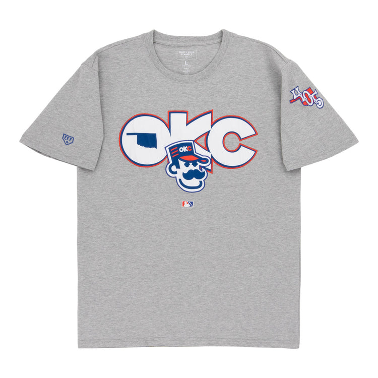 The Ebbets Field Oklahoma City 89Ers MiLB Fashion Cotton Short Sleeve Tee is a heather gray T-shirt with a large logo and cartoon character on the chest, plus 405 patches and other symbols on the sleeves.