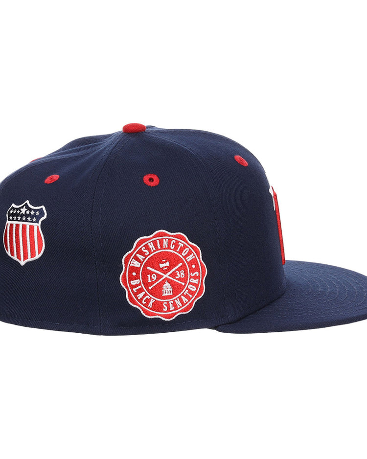 The Ebbets Field Washington Black Senators NLB Varsity Fitted Ballcap is a navy blue cap with a flat brim, detailed with red and white embroidered shield and circular Washington Senators logo patches on the side.