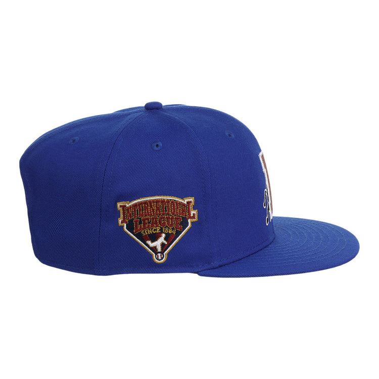 The Ebbets Field Durham Bulls MiLB Snapback Ballcap is a blue flat-brim hat with an embroidered International League Since 1884 patch on the left side, shown from a left-side angle.