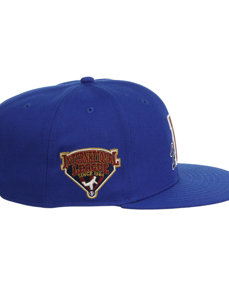 The Ebbets Field Durham Bulls MiLB Snapback Ballcap is a blue flat-brim hat with an embroidered International League Since 1884 patch on the left side, shown from a left-side angle.