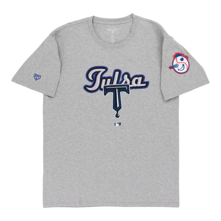 The Ebbets Field Tulsa Drillers MiLB Fashion Cotton Short Sleeve Tee is a gray shirt with “Tulsa” in blue and white script and a large T on the front. The left sleeve features the Drillers’ baseball mascot head with a red feather headband.