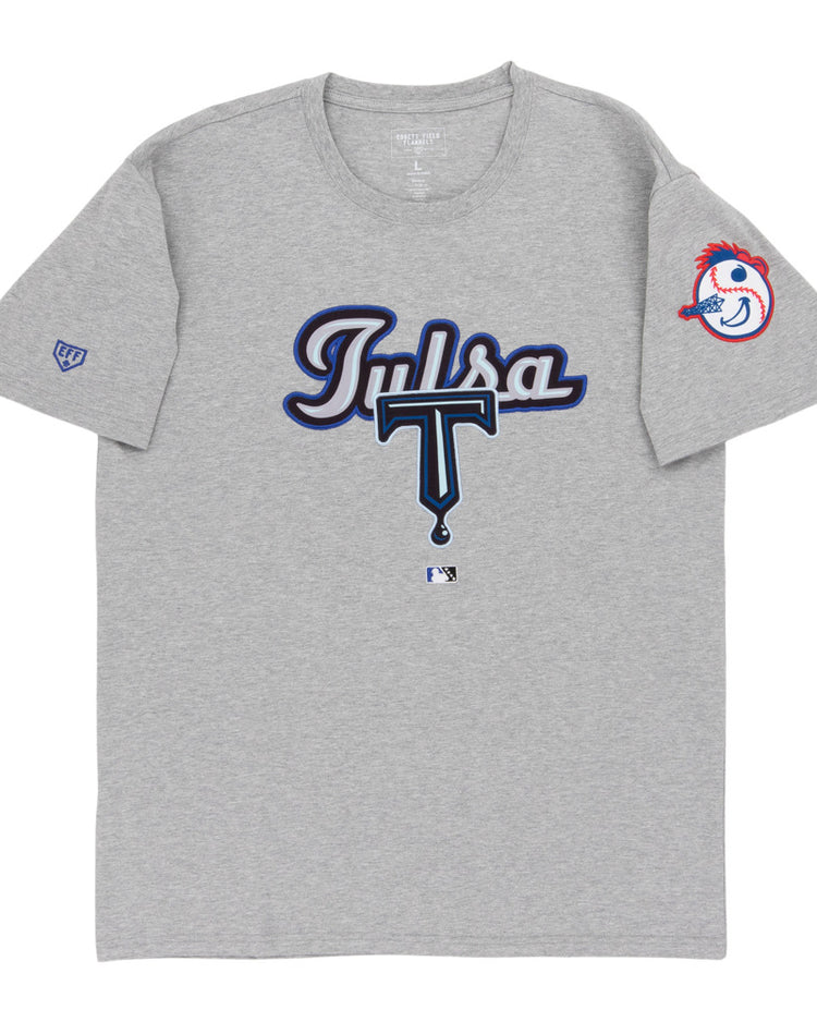 The Ebbets Field Tulsa Drillers MiLB Fashion Cotton Short Sleeve Tee is a gray shirt with “Tulsa” in blue and white script and a large T on the front. The left sleeve features the Drillers’ baseball mascot head with a red feather headband.