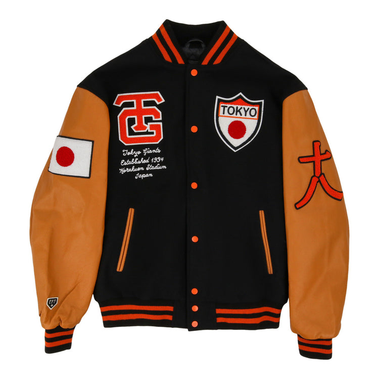 The Ebbets Field Tokyo Giants Dna Varsity Jacket is black with tan sleeves, orange accents, a Japanese flag patch on the right sleeve, and embroidered TOKYO shield, script, and letters on the chest.