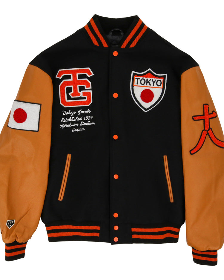The Ebbets Field Tokyo Giants Dna Varsity Jacket is black with tan sleeves, orange accents, a Japanese flag patch on the right sleeve, and embroidered TOKYO shield, script, and letters on the chest.