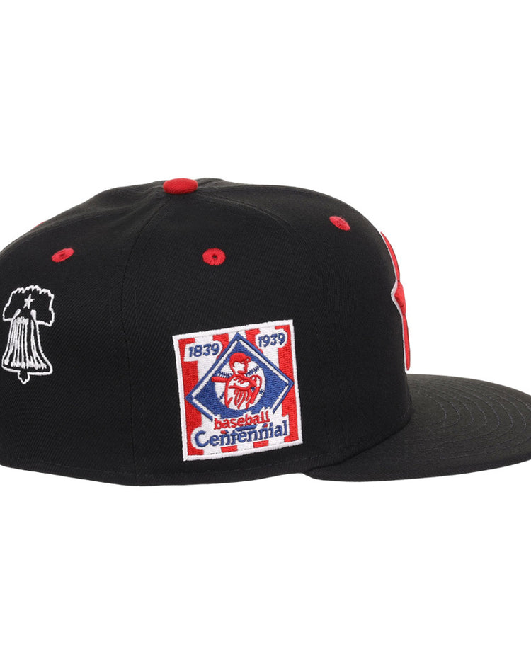 The Ebbets Field Philadelphia Stars NLB Varsity Fitted Ballcap is a black cap with red accents, flat brim, vent holes, and side embroidered patches—a Baseball Centennial 1839-1939 logo and a white outlined bell.
