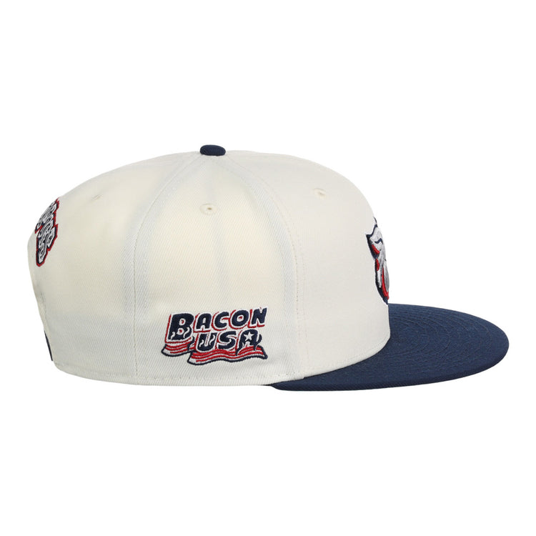 The Lehigh Valley Ironpigs MiLB Snapback Ballcap by Ebbets Field is a white cap with a navy brim, embroidered Bacon USA text and flag on the side, plus additional patches on the front and back.