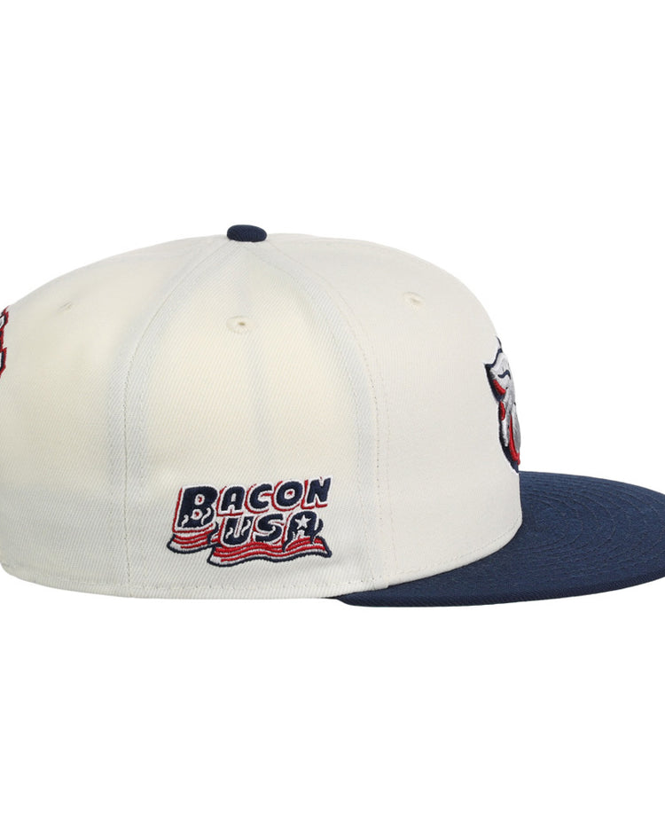 The Lehigh Valley Ironpigs MiLB Snapback Ballcap by Ebbets Field is a white cap with a navy brim, embroidered Bacon USA text and flag on the side, plus additional patches on the front and back.