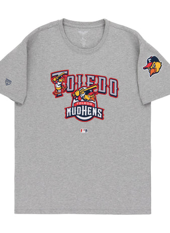 The Ebbets Field Toledo Mud Hens MiLB Fashion Cotton Short Sleeve Tee is gray, with the team’s logo and name in red, gold, and white on the chest, plus a Mud Hens logo patch on the left sleeve.