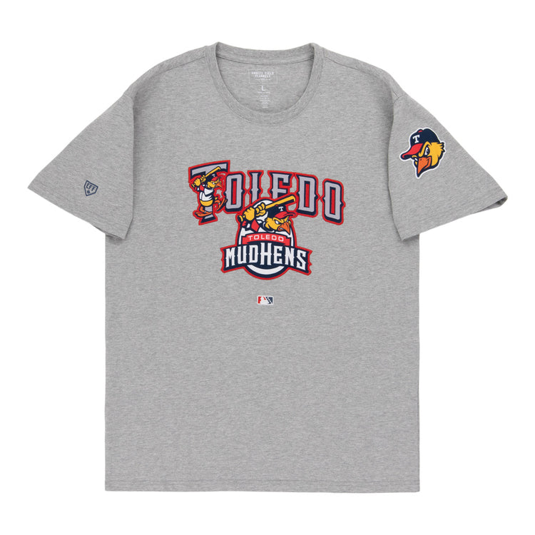 The Ebbets Field Toledo Mud Hens MiLB Fashion Cotton Short Sleeve Tee is gray, with the team’s logo and name in red, gold, and white on the chest, plus a Mud Hens logo patch on the left sleeve.