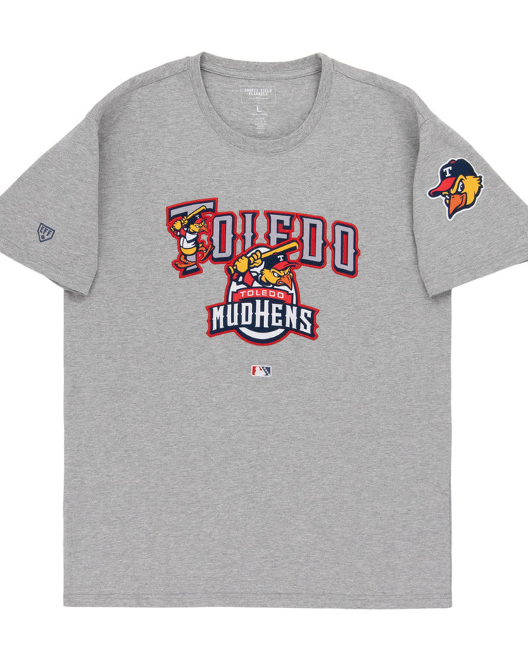The Ebbets Field Toledo Mud Hens MiLB Fashion Cotton Short Sleeve Tee is gray, with the team’s logo and name in red, gold, and white on the chest, plus a Mud Hens logo patch on the left sleeve.