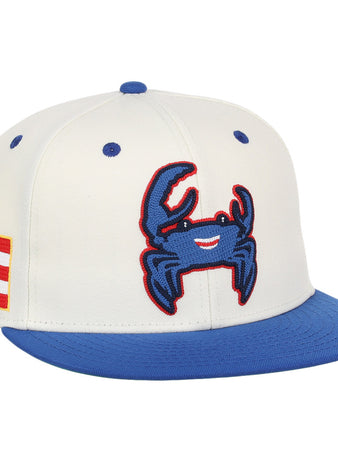 The Ebbets Field Santurce Cangrejeros Chainstitch Fitted Ballcap is a white cap with a blue brim, featuring a smiling blue crab patch on the front and a Puerto Rican flag patch on the side.